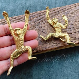 May include: Two gold monkey shaped hooks on a wooden surface. The hooks are in a playful pose with their arms raised.