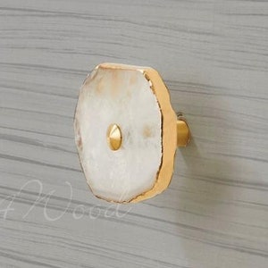 Pode incluir: Dois puxadores de armário com um design branco e dourado. Os puxadores têm uma forma natural e irregular com uma borda dourada e um parafuso dourado. Os puxadores são montados em uma superfície de madeira cinza claro.