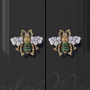 Può includere: Due pomelli per armadi a forma di ape dorati con ali verdi e dettagli di perle bianche. I pomelli sono montati su una porta nera.