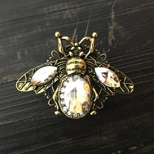 Könnte beinhalten: Eine goldfarbene Bienenbrosche mit klaren Kristall-Akzenten. Die Biene hat detaillierte Flügel und einen Körper mit einem Kristall-Mittelpunkt.