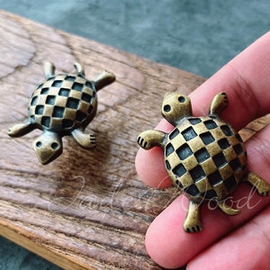 Op de afbeelding: Twee kleine, bronzen schildpadbeeldjes. De schildpadden hebben een dambordpatroon op hun schild en zijn geplaatst op een houten oppervlak. De schildpadden zijn gedetailleerd met zichtbare poten, koppen en staarten.