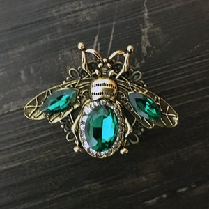 Könnte beinhalten: Eine goldfarbene Bienenbrosche mit grünen Kristall-Akzenten. Die Biene hat ein detailliertes Design und ist vor einem dunklen Holzhintergrund platziert.