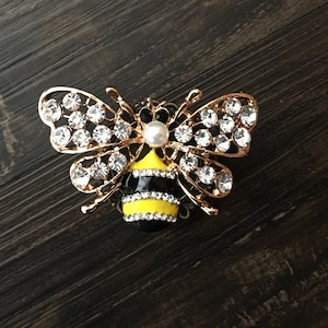 May include: A gold bee brooch with black and yellow stripes and clear rhinestones. The bee has a pearl in the center of its head.