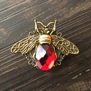 Op de afbeelding: Een gouden bij broche met een rode facetgeslepen edelsteen. De bij heeft ingewikkelde details en een delicaat ontwerp.