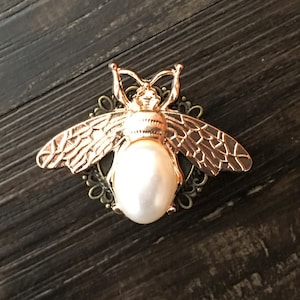 Könnte beinhalten: Eine goldene Bienenbrosche mit einem Perlenmittelpunkt. Die Biene hat detaillierte Flügel und ein zartes Blumenmuster.
