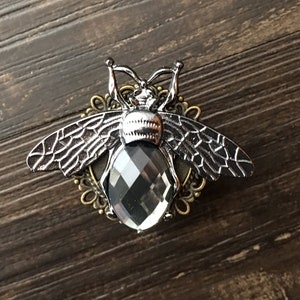 May include: A detailed bee brooch with a faceted, dark gray gemstone body and silver-toned wings. The brooch has an ornate, bronze-colored filigree base, creating a striking contrast against the dark wood grain background.