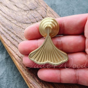 Puede incluir: Un pomo de armario de metal dorado con un diseño en forma de abanico. El pomo se sostiene en una mano.