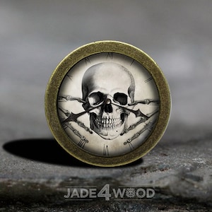 Puede incluir: Una esfera de reloj redonda de color dorado antiguo con números romanos. La esfera del reloj presenta una ilustración en blanco y negro de una calavera con huesos cruzados.