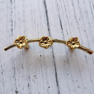 May include: Gold-toned metal cabinet hardware with a floral design. The hardware features three stylized flowers on a curved branch.