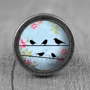 May include: A round, silver knob with a blue background and a floral pattern. Five black birds are perched on a wire in front of the flowers.