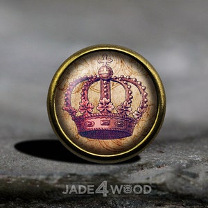 Può includere: Un bottone rotondo dorato con un'immagine in stile vintage di una corona rosa e viola con una croce in cima. La corona è circondata da un motivo floreale decorativo.