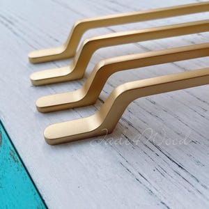 以下が含まれることがあります： 明るい色の木材の表面に斜めに配置された4つの金色のキャビネットプル。各プルは長方形のベースと湾曲したハンドルを備えています。プルは金属製で、マット仕上げです。背景は、水色と白の木材の表面です。