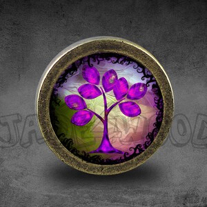 Puede incluir: Un colgante redondo de color bronce, con un diseño de árbol morado estilizado. El árbol tiene un tronco morado oscuro y hojas moradas vibrantes. El fondo tiene una mezcla de colores, incluyendo verde, rosa y blanco, con un borde decorativo negro.