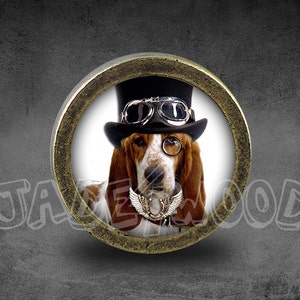 May include: A round, brass-colored, decorative knob with a picture of a basset hound wearing a top hat and goggles. The dog is looking directly at the camera.