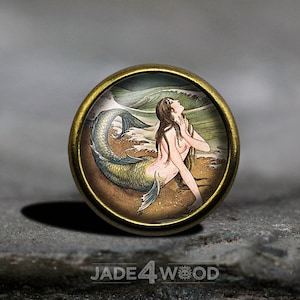 May include: A round, brass-coloured button featuring a vintage illustration of a mermaid with a green tail, sitting on a sandy beach with waves behind her.
