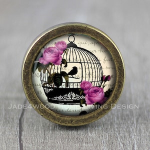 Puede incluir: Un pomo de armario de estilo vintage con una base redonda de color latón y una parte superior de vidrio que presenta una jaula de pájaros negra con rosas rosas y un pájaro negro en su interior. La parte superior de vidrio tiene un fondo blanco con texto negro.