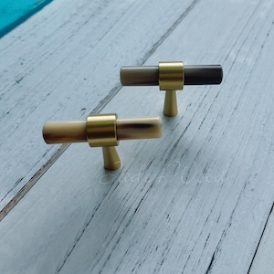 Könnte beinhalten: Zwei Schrankgriffe mit zylindrischem Design. Die Griffe haben eine hellbraune und dunkelbraune Oberfläche mit goldfarbenen Metallakzenten. Die Griffe sind auf einer hellblauen und weißen Holzoberfläche angeordnet.