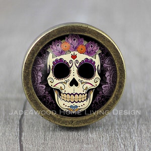 May include: A cabinet knob with a Day of the Dead sugar skull design. The skull is cream-coloured with black and purple details, a floral crown, and a heart. The knob is set in a bronze-coloured metal frame. The design is circular.