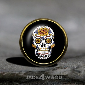 May include: A round, gold-colored metal pin with a black background and a white sugar skull with colorful floral details. The skull has a yellow rose on top of its head and yellow, blue, and orange flowers around its eyes.