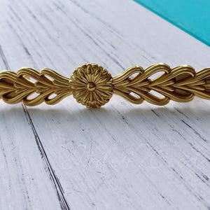 以下が含まれることがあります： 装飾的な花のデザインが施された金色のキャビネットの取っ手。取っ手は円形のセンターピースと花の模様を持っています。