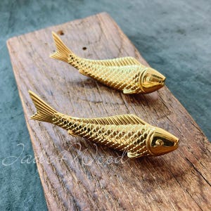 Può includere: Due tiretti per cassetti o ante a forma di pesce color oro. I tiretti sono su una superficie di legno.