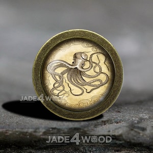May include: A round, brass-colored button with a vintage illustration of an octopus on a cream-colored background. The octopus has eight tentacles and is surrounded by a decorative border.