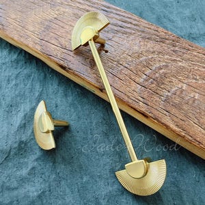 Op de afbeelding: Twee gouden metalen kastgrepen met een waaiervormig ontwerp. De grotere greep heeft een lange, rechte stang die de twee waaiervormige delen verbindt. De kleinere greep is een enkel waaiervormig deel.