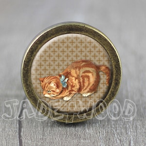May include: A round, brass-coloured knob with a vintage-style image of a brown and orange tabby cat wearing a blue bow. The cat is looking to the left and the background is a brown and white patterned design.