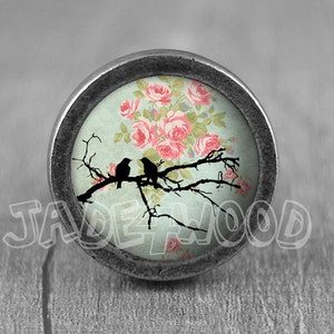 May include: A silver knob with a glass insert featuring a vintage floral design with pink roses and two black birds perched on a branch.