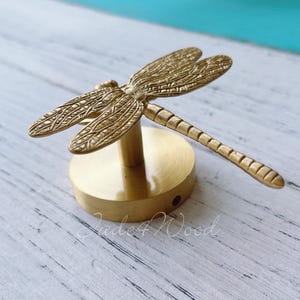 Könnte beinhalten: Eine Messing-Libellenskulptur mit detaillierten Flügeln und einem segmentierten Schwanz. Die Libelle ist auf einem runden Sockel montiert. Die Skulptur ist ein dekoratives Akzentstück.