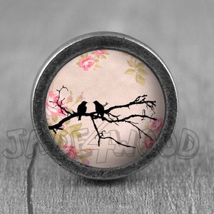 May include: A silver knob with a round, glass top featuring a pink floral pattern and two black birds perched on a branch.