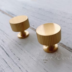 May include: Two gold-colored cabinet knobs with a cylindrical design and vertical grooves. The knobs have a circular base and a flat top. The knobs are made of metal and are set against a light-colored wooden surface.