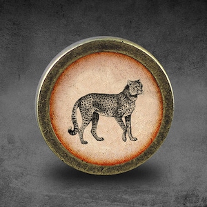 Puede incluir: Un pomo redondo de color latón con un círculo elevado de color naranja-marrón en el centro. El círculo presenta una ilustración en blanco y negro de un guepardo.
