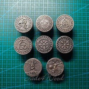 May include: Eight circular silver-toned metal tokens featuring embossed designs including a triskelion, a triquetra, a valknut, a vegvisir, and trees of life, all arranged in a grid pattern on a green grid mat.