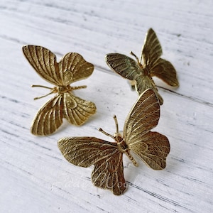Puede incluir: Tres adornos de mariposa de metal de color dorado. Las mariposas tienen alas detalladas y están dispuestas en un grupo sobre una superficie de madera blanca.