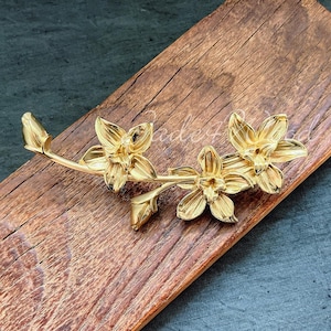 Peut inclure: Une broche en métal doré représentant trois fleurs d'orchidées stylisées avec une tige et des feuilles. La broche est posée sur une surface en bois.