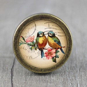 Peut inclure: Un bouton de style vintage avec une finition en laiton présente un motif circulaire avec deux oiseaux bleus perchés sur une branche avec des fleurs roses. L'arrière-plan est une carte postale de couleur crème avec le texte "MAS.  N.  MILN.  309.  SH."