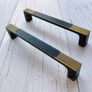 Puede incluir: Dos tiradores de armario negros con detalles dorados. Los tiradores tienen forma rectangular con la parte superior e inferior ligeramente curvadas. Los detalles dorados son en forma de líneas que recorren la longitud del tirador.