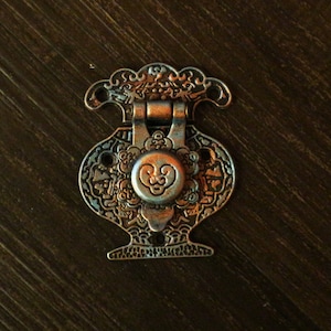 Puede incluir: Un cierre de metal adornado en forma de jarrón con intrincados diseños florales. El cierre tiene un pomo en forma de corazón con un diseño estilizado.