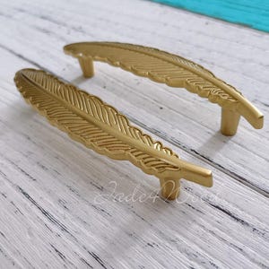 May include: Two gold-toned feather-shaped cabinet pulls. The pulls are made of metal and have a detailed feather design.