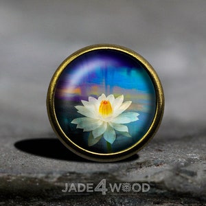 May include: A white water lily with a yellow centre is depicted in a glass cabochon, set in a brass setting. The background is a blue and purple watercolour wash.