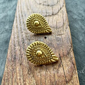 Puede incluir: Un par de pendientes de forma de lágrima de color dorado con un diseño texturizado y adornado. Los pendientes se muestran sobre una superficie de madera.
