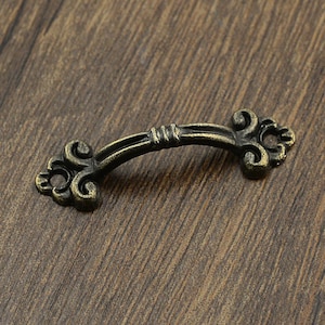 Könnte beinhalten: Ein kleiner, antiker, bronzefarbener Metall-Schubladengriff mit einem dekorativen, verzierten Design. Der Griff hat eine gebogene Form und zwei Enden in Form von Schnörkeln.