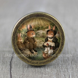 Puede incluir: Un pomo de armario redondo de color latón con un inserto de vidrio que presenta una ilustración caprichosa de tres conejos en un entorno de bosque. Los conejos llevan ropa y uno de ellos sostiene un pequeño barco.