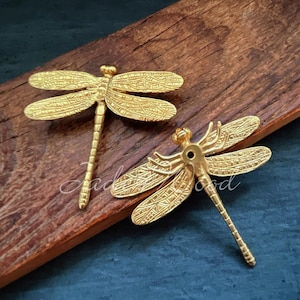 May include: Two gold-toned dragonfly drawer pulls with intricate details. The dragonfly pulls have a hole in the centre for mounting.