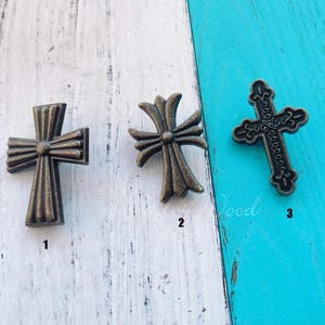 May include: Three bronze-coloured cross-shaped decorative embellishments. The crosses have varying designs, including a ribbed, a floral, and a detailed filigree style. They are arranged on a white and turquoise wooden surface.