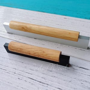 May include: Two contemporary cabinet pulls. One is white with a light wood handle, and the other is black with a light wood handle. The handles are cylindrical and made of light-coloured wood. The pulls are set against a light wood background.