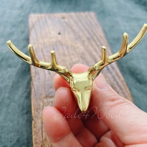 Può includere: Ganci da parete in metallo dorato a forma di testa di cervo su sfondo di legno rustico. Il gancio ha un design a tre punte, che ricorda le corna.