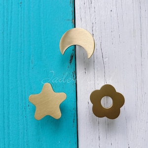 Könnte beinhalten: Drei goldene Metall-Schubladengriffe in Form einer Mondsichel, eines Sterns und einer Blume. Die Griffe befinden sich auf einem blau-weißen Holzhintergrund.