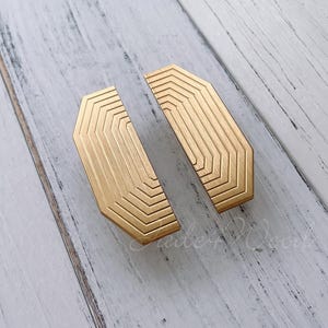 May include: Two gold-toned metal cabinet pulls with a geometric design. The pulls are shaped like octagons with rounded corners.